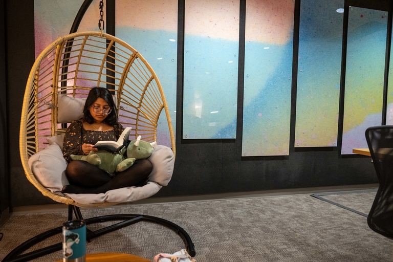 Student lounging in a suspended egg chair while reading a book. A weighted stuffed animal is in her lap, and paneled, colorful artwork is in the background. 
