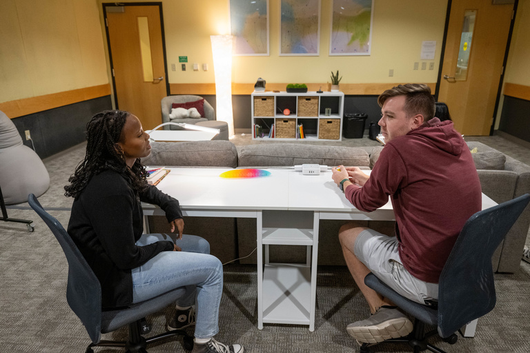 Male and female student speaking at a desk, with a background showing warm lighting, artwork, and a storage cube with fidget toys