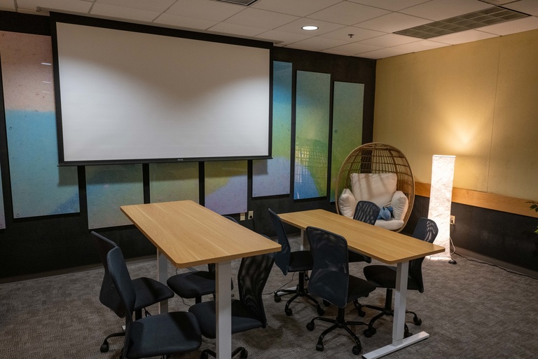 Two tables, one standing height, and the other sitting height, surrounded by height-adjustable swivel chairs. An egg chair and warm light is in the background. The back wall shows a projector screen 