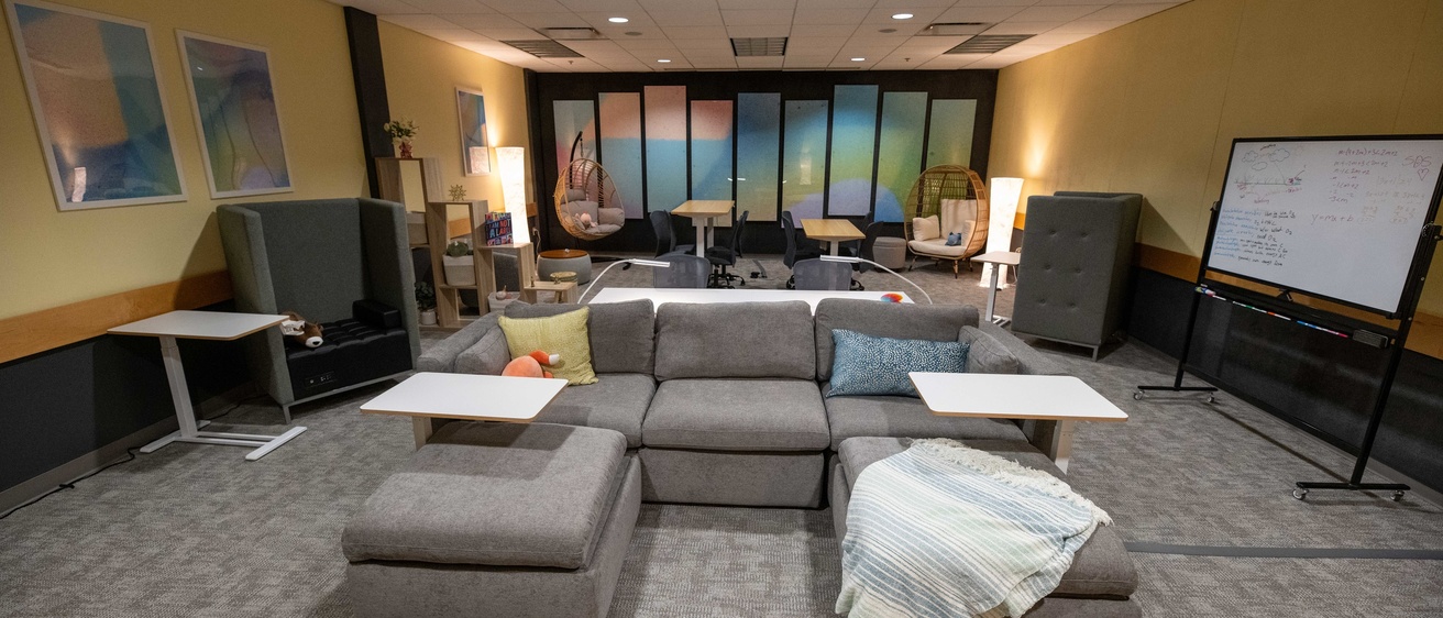 SDS Lounge. Big grey couch, with whiteboard in the background with students' work. Yellow walls and warm lighting, with gradient artwork lining the walls. There are highback chairs, and height-adjustable workstations. 