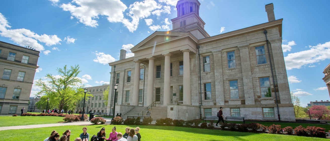 The Old Capitol building in front of a bright blue sky, with several people sitting in a circle in the grass in front of it.