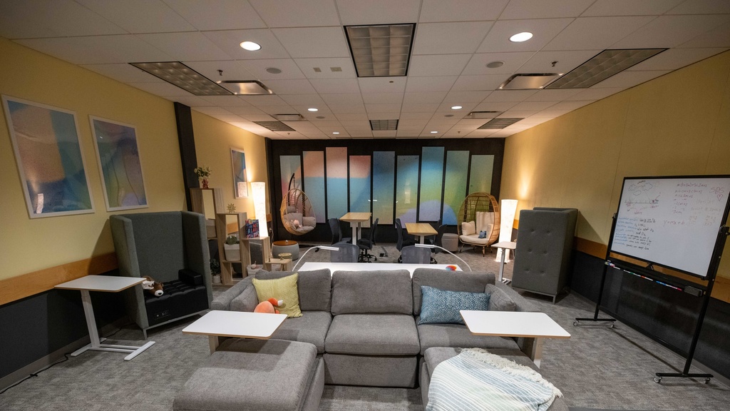The interior of the SDS Lounge. There is a big comfy couch with pillows and weighted stuffed animals. There is a whiteboard, along with basket chairs, high back chairs, and height adjustable work stations. There are warm light lamps lighting up the gradient artwork on the back and left yellow walls. 